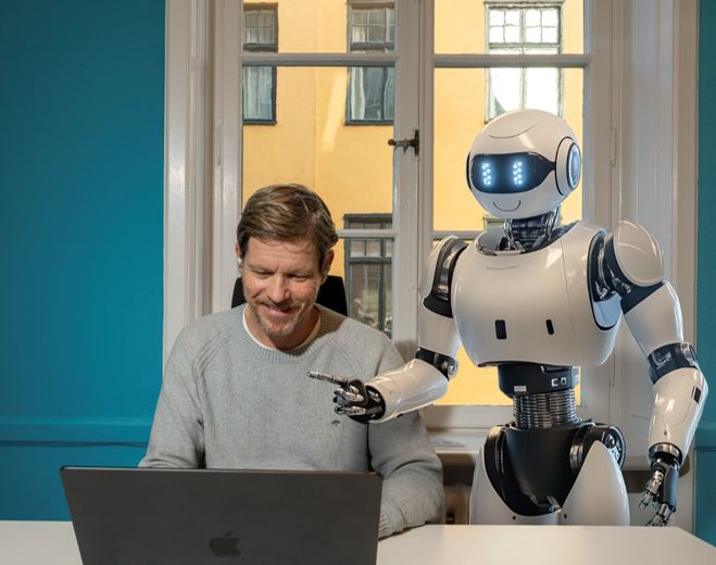 En man sitter vid en bärbar dator och ler medan en vit och blå robot pekar mot skärmen, i ett rum med turkosa väggar.