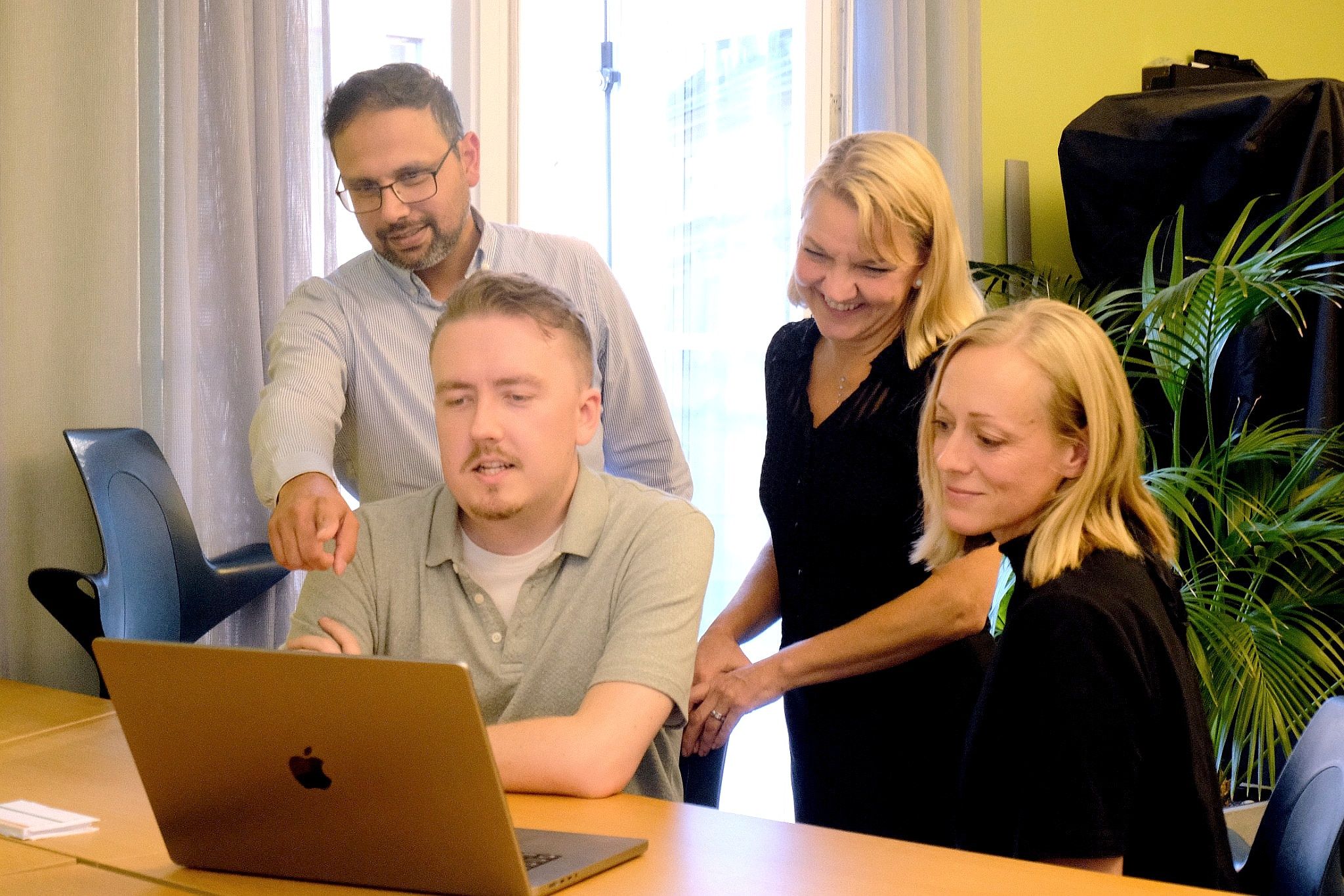 Fyra kollegor samlade kring en bärbar dator i ett kontorsrum med gul vägg; en man pekar mot skärmen medan de andra ler.