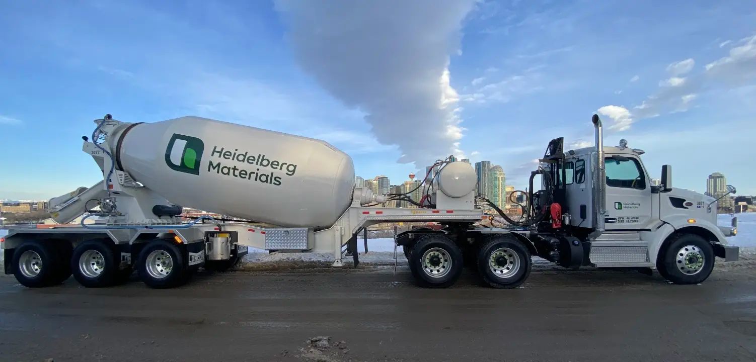 En cementblandarbil märkt "Heidelberg Materials" står parkerad på en snökantad väg med en stadssilhuett i bakgrunden under en blå himmel.