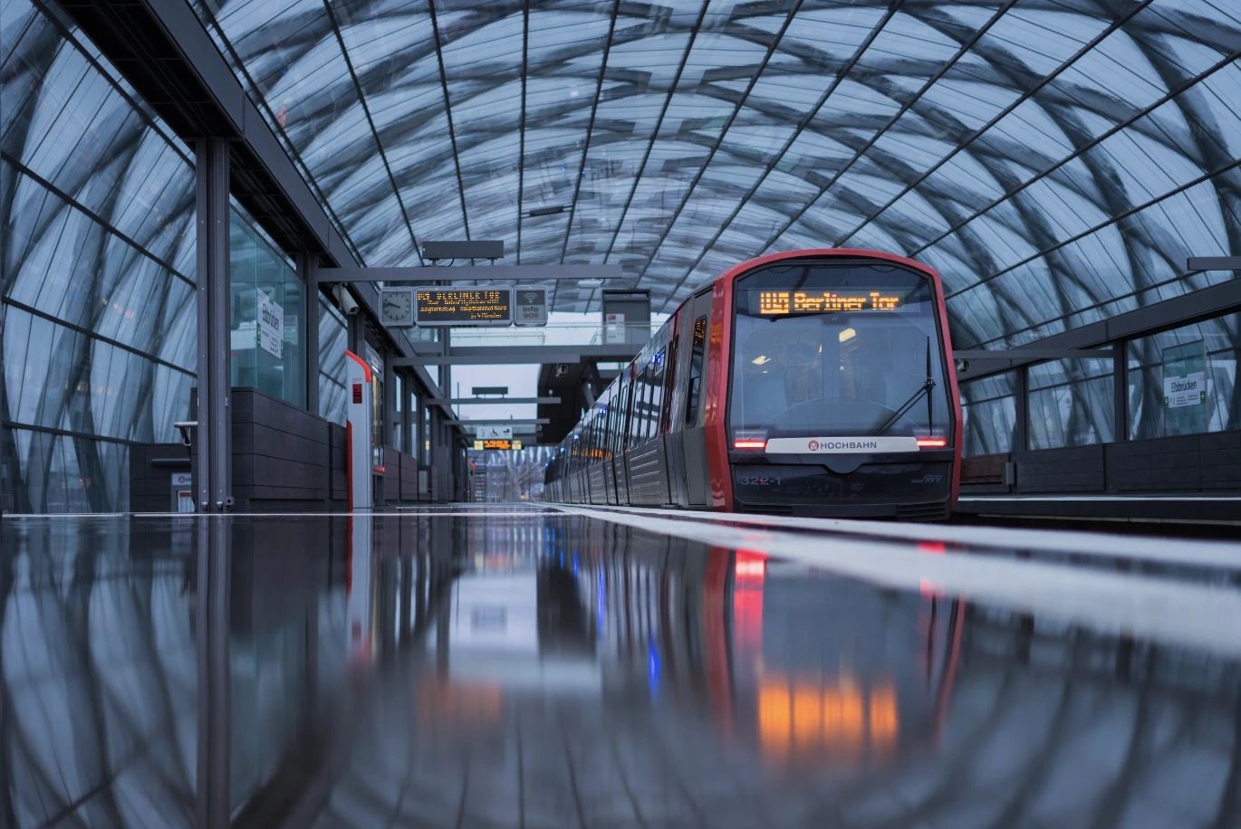 Ett rött tåg märkt "Berliner Tor" är stationerat på en reflekterande plattform under ett välvt glastak. Skyltar ovanför visar "Bahrenfeld", "Elbbrücken" och annan stationsinformation.