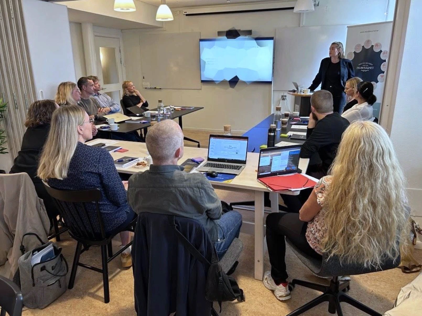 Människor deltar i ett mötesrum och lyssnar på en högtalare nära en skärm som visar innehåll. Bärbara datorer och anteckningsblock ligger på borden och en whiteboardtavla syns i bakgrunden.