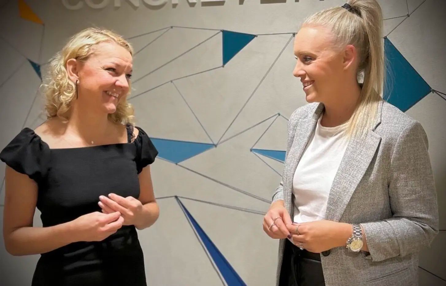 Två kvinnor ler och är engagerade i konversation, står framför en vägg med en geometrisk design. En kvinna bär en svart klänning, den andra en grå kavaj.